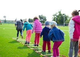 Children enjoy an outdoor activity on a grassy field, stepping over a ladder.